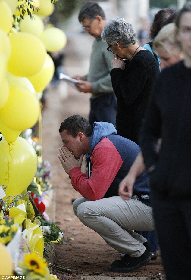 Aaron Leeson-Woolley (crouching) accompanies Stephanie Scott's mother (right) and father Robert (at rear) to look at floral tributes laid to Ms Scott outside Leeton High School  