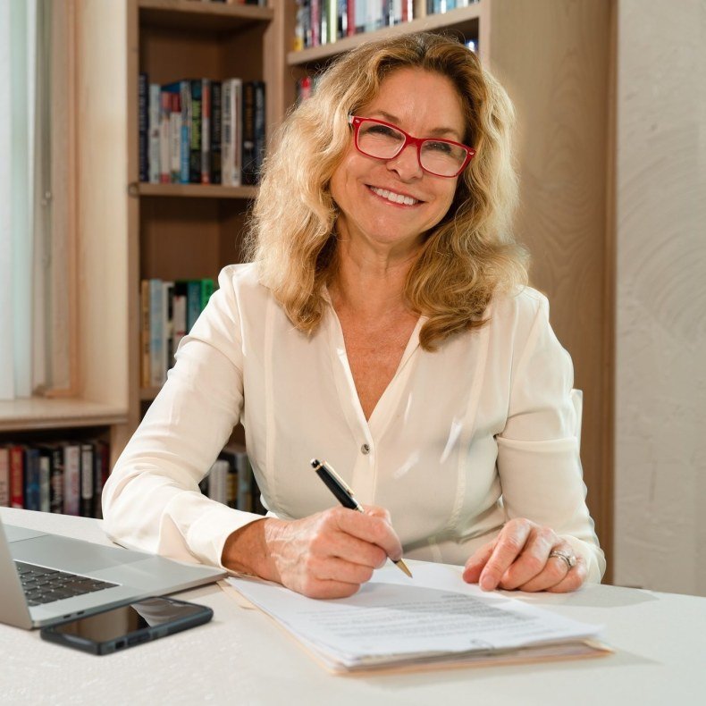 A woman in red glasses and a white blouse sits at a desk with a laptop, holding a pen over a piece of paper, smiling at the camera. 