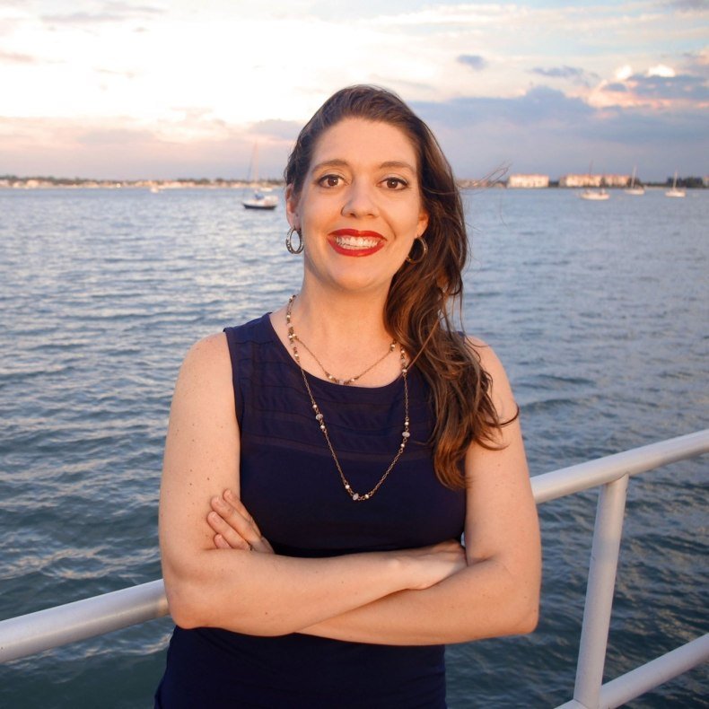 A woman in a sleeveless dress with arms crossed stands smiling at the camera in front of the water. 